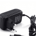 Um adaptador de energia ALIMENTADOR 12V 1A C/ FICHA 5,5X2,1MM com plugue europeu e um cabo que termina em um conector cilíndrico de 5,5x2,1mm está sobre uma superfície branca. O adaptador preto apresenta texto e símbolos na lateral, e o cabo é bem enrolado e preso com um laço preto.